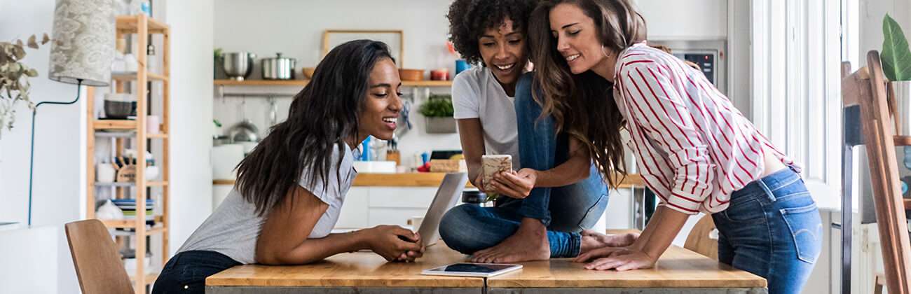 Three women with multiple mobile devices gather around a table looking at a mobile phone